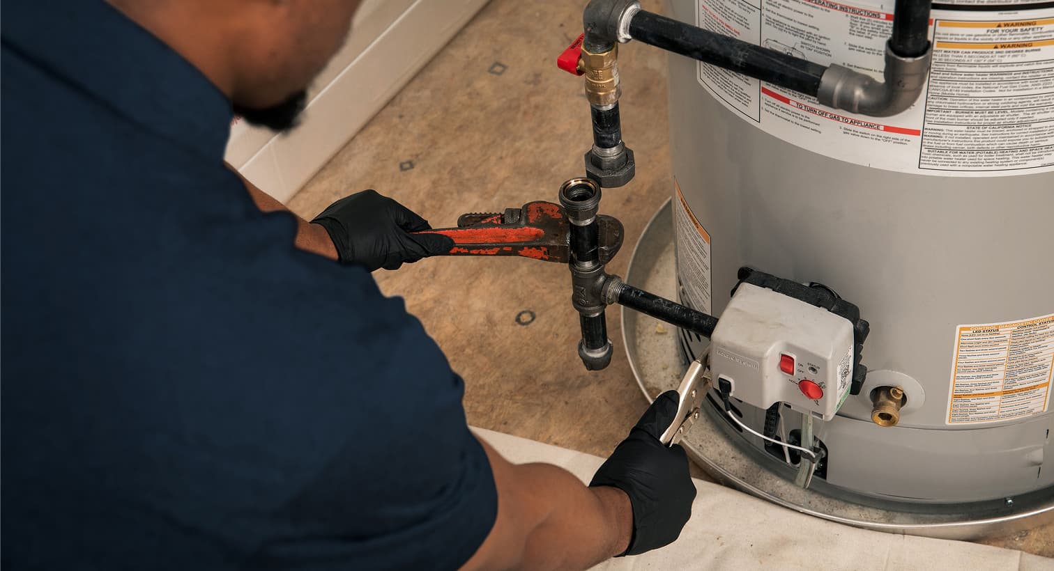 An Elite plumber wearing black gloves uses a pipe wrench to repair the gas line and control valve assembly on a residential water heater tank.