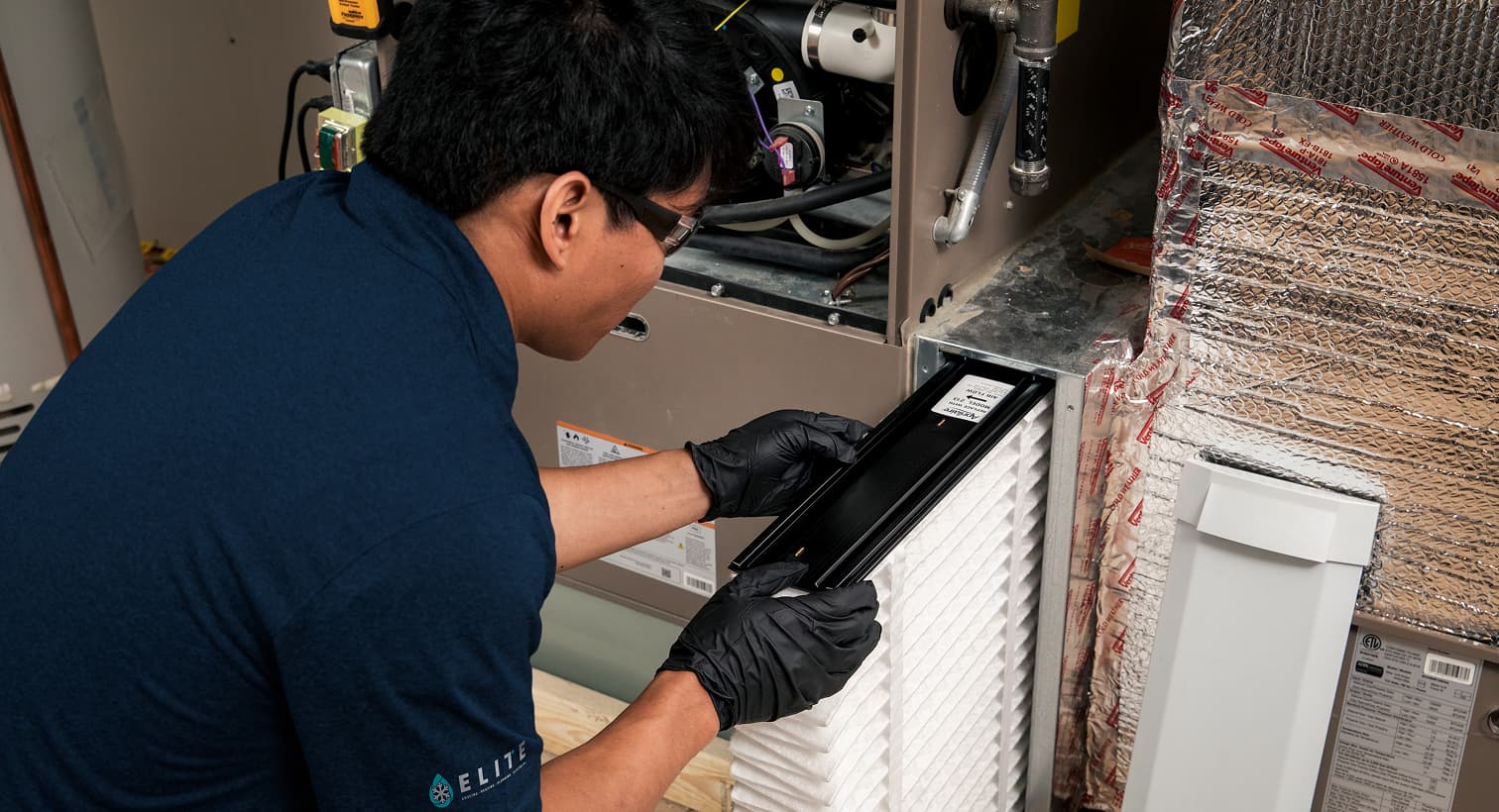 A professional Elite HVAC technician replacing a clean pleated air filter in an indoor air handler unit during a routine AC maintenance tune-up.