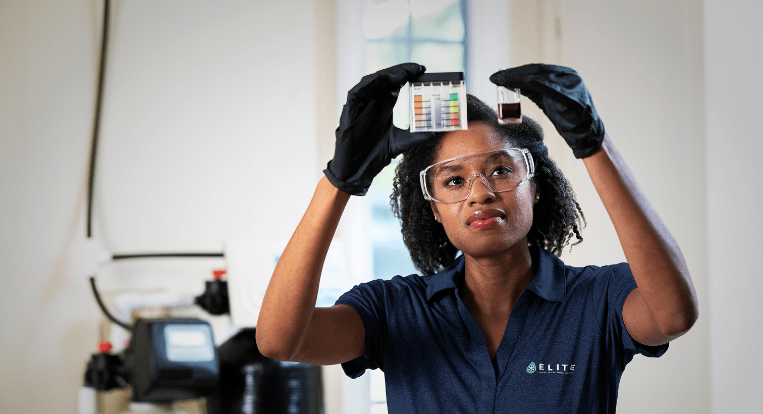 A Elite technician in a blue uniform and safety gear compares a water sample to a testing chart in front of a water softener system.